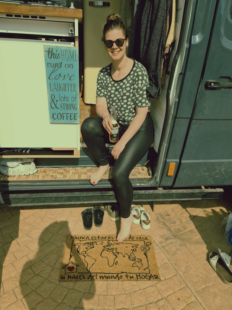 A woman sitting on the step of her campervan holding a bottle of beer. The sign next to her reads 'this home runs on love, laughter & lots of strong coffee'