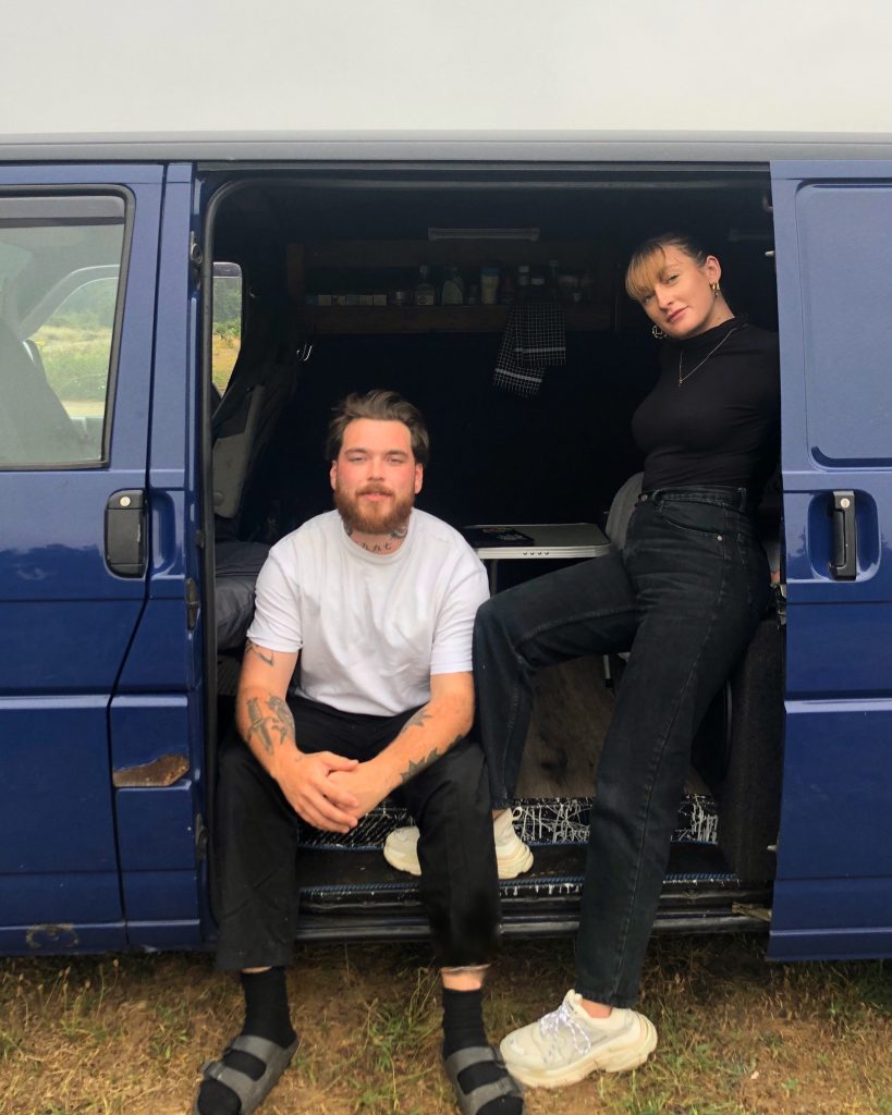 Blue T4 campervan with the sliding door open. Billy is sat on the floor with his feet out while Leanne is sat higher up inside the van.
