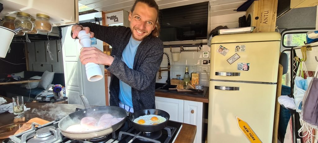 A man cracking some fresh pepper over bacon in a pan whilst frying some eggs in the pan next to it