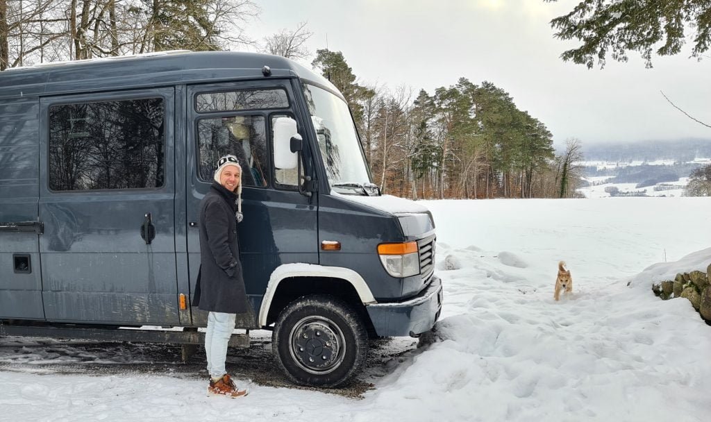 Man standing smiling beside his campervan in the snow whilst a small dog runs toward him.