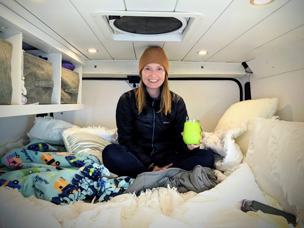 Girl sitting cross legged in her campervan. The walls are white and she is sat on the bed with a green cup in her hand