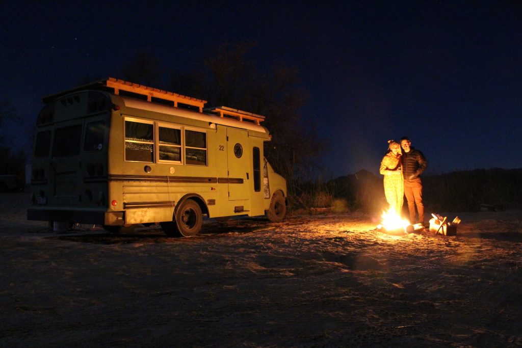The bus at night time. A campfire outside with Andy and Ayana stood in front of it