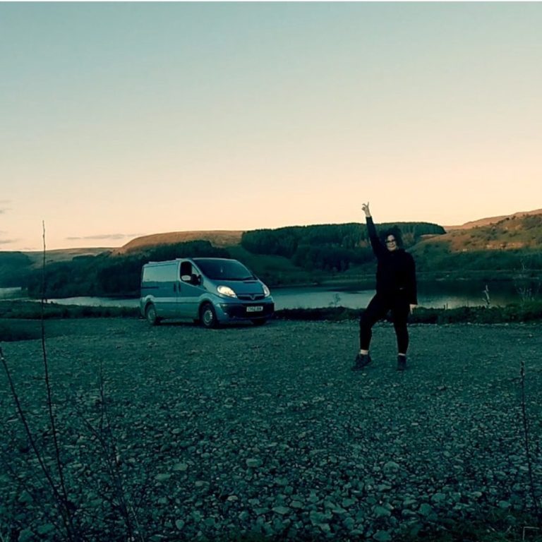 Miss Kiff is stood with one arm up in the air. She is dressed in all black. Her van is behind her with the sun setting in the background